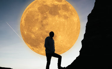 A person climbing a rocky hillside is silhouetted against a golden full moon with crater details.