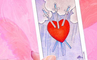 A hand holds a Three of Swords Tarot card, featuring a red heart pierced with three swords.