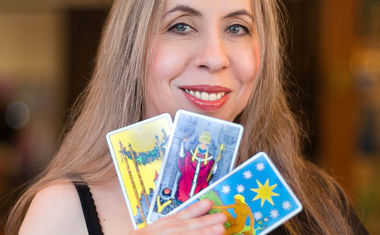 A photograph of Tarot reader Theresa Reed, a white woman with long hair holding three Tarot cards.