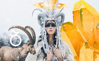 A woman with a horned headdress and dark makeup stands in the snow beside a goat.