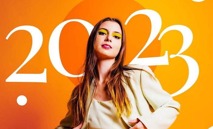 A woman wearing a white suit and orange eyeshadow stands in front a backdrop reading "2023."