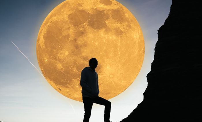 A person climbing a rocky hillside is silhouetted against a golden full moon with crater details.