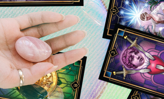 A hand holding a polished rose quartz stone hovers over a table full of Tarot cards.