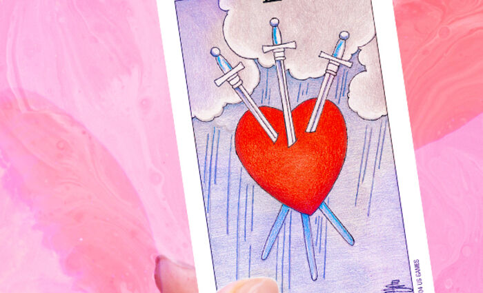A hand holds a Three of Swords Tarot card, featuring a red heart pierced with three swords.